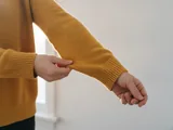 Hände prüfen die Ärmellänge einer senfgelben Strickjacke vor einer weißen Wand
