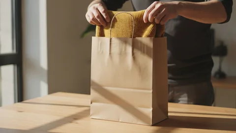 Hände legen einen gefalteten senfgelben Pullover in eine schlichte Papiertüte